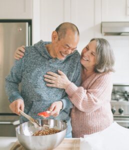 Smiling senior couple cooking in a kitchen. The woman is standing next to the man with her arms around him. The man is hold cooking utensils over a bowl of food.
